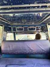 The interior of a vehicle featuring a blue, patterned ceiling with circular designs. There is a rear view of a single seat covered in a textured, purple material. A decorative item hangs from the vehicle's ceiling. The partial view of a driver's head is visible beyond the seat, with part of the road ahead visible through the windshield.