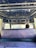 The interior of a vehicle featuring a blue, patterned ceiling with circular designs. There is a rear view of a single seat covered in a textured, purple material. A decorative item hangs from the vehicle's ceiling. The partial view of a driver's head is visible beyond the seat, with part of the road ahead visible through the windshield.