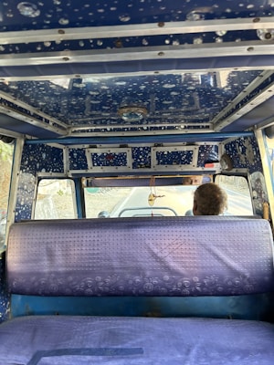 The interior of a vehicle featuring a blue, patterned ceiling with circular designs. There is a rear view of a single seat covered in a textured, purple material. A decorative item hangs from the vehicle's ceiling. The partial view of a driver's head is visible beyond the seat, with part of the road ahead visible through the windshield.