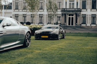 Two luxury cars are parked on a well-manicured lawn in front of an elegant, large house with multiple windows and shutters. The car in the foreground is a silver sports car, and the one in the background is a black sports car with a visible license plate. The setting includes trimmed hedges and a couple of slender trees.