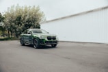 A green SUV is parked on a paved surface in front of a large white building with a vertical panel design. Trees with dense foliage are visible to the left, suggesting a mix of urban and natural surroundings.