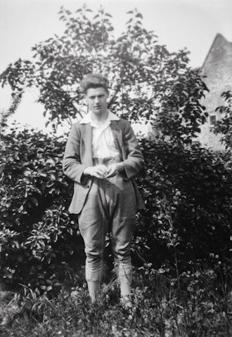 A young person stands in front of lush shrubbery, dressed in a suit with a white shirt. The expression is neutral, and there is an old stone building partially visible in the background.