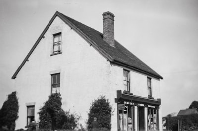 A two-story building with a brick chimney and a steep roof. The structure is housing a small grocery store with signs reading 'Grocery' and 'Provisions' above the windows. The exterior is plain with a couple of windows visible and some small bushes surrounding the building.