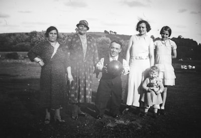 A group of six people, consisting of four adults and two children, stand outdoors on grass against a backdrop of trees. The adults are dressed in clothing typical of early to mid-20th century, and one man holds a ball. The children, one with a doll, are positioned in front of the adults. The scene conveys a sense of a family or community gathering during daylight hours.