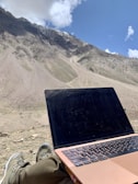 A person is seated outdoors with a laptop on their lap, looking towards a vast, barren mountainous landscape under a bright blue sky partially covered by clouds. The person is likely enjoying the view while working or relaxing.