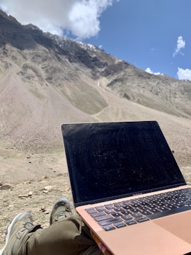A person is seated outdoors with a laptop on their lap, looking towards a vast, barren mountainous landscape under a bright blue sky partially covered by clouds. The person is likely enjoying the view while working or relaxing.