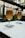 Two tall glasses of beer are placed on a table with branded coasters in an outdoor setting. The background features blurred views of a patio with greenery and a building facade.