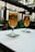 Two tall glasses of beer are placed on a table with branded coasters in an outdoor setting. The background features blurred views of a patio with greenery and a building facade.