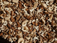A close-up view of a mixture of various nuts including almonds, cashews, and walnuts. The nuts are spread out, showcasing their natural textures and colors, with a mix of light and dark browns and creamy whites.