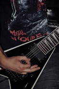 A person's hand is playing a black electric guitar with a V-shaped body. The person is wearing a black t-shirt with a graphic design featuring red lettering and imagery. The overall setting conveys a dark, intense atmosphere typical of metal music.
