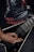 A person's hand is playing a black electric guitar with a V-shaped body. The person is wearing a black t-shirt with a graphic design featuring red lettering and imagery. The overall setting conveys a dark, intense atmosphere typical of metal music.