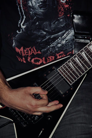 A person's hand is playing a black electric guitar with a V-shaped body. The person is wearing a black t-shirt with a graphic design featuring red lettering and imagery. The overall setting conveys a dark, intense atmosphere typical of metal music.