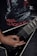 A person's hand is playing a black electric guitar with a V-shaped body. The person is wearing a black t-shirt with a graphic design featuring red lettering and imagery. The overall setting conveys a dark, intense atmosphere typical of metal music.