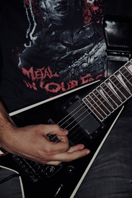 A person's hand is playing a black electric guitar with a V-shaped body. The person is wearing a black t-shirt with a graphic design featuring red lettering and imagery. The overall setting conveys a dark, intense atmosphere typical of metal music.