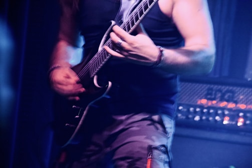 A person wearing a sleeveless top and camouflage pants plays an electric guitar with visible intensity. The atmosphere is dimly lit, with a focus on the person's muscular arms and the guitar. Amplifiers in the background suggest a live performance setting.