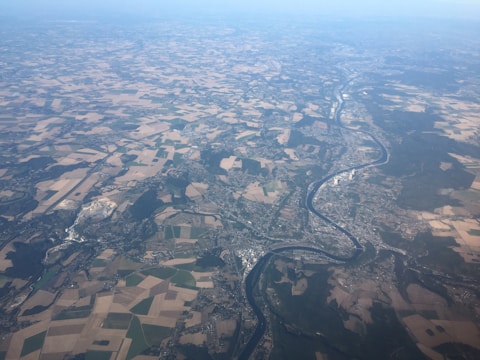 An aerial view of a landscape with a winding river cutting through farmland and urban areas. The fields are various shades of green and brown, indicating a patchwork of different crops or land uses. Urban areas with clusters of buildings are visible alongside the river. The horizon fades into a hazy blue sky.