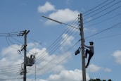 A person is climbing a utility pole, equipped with a harness and tools, to work on electrical wires. Another utility pole with more complex wiring and equipment is seen nearby. The background features a clear blue sky with a few scattered clouds.