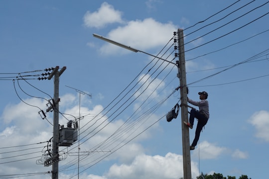 A person is climbing a utility pole, equipped with a harness and tools, to work on electrical wires. Another utility pole with more complex wiring and equipment is seen nearby. The background features a clear blue sky with a few scattered clouds.