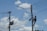 A person is climbing a utility pole, equipped with a harness and tools, to work on electrical wires. Another utility pole with more complex wiring and equipment is seen nearby. The background features a clear blue sky with a few scattered clouds.