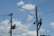 A person is climbing a utility pole, equipped with a harness and tools, to work on electrical wires. Another utility pole with more complex wiring and equipment is seen nearby. The background features a clear blue sky with a few scattered clouds.