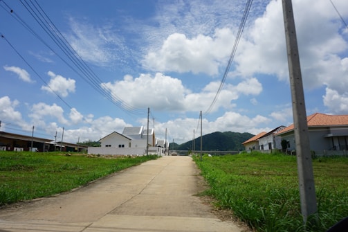 A paved road leads through a residential area with grassy lawns on either side. White houses with red roofs are visible, and utility poles with power lines line the road. In the distance, a mountain range is visible under a partly cloudy sky.