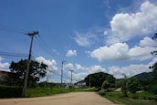Power lines stretch across a clear blue sky with fluffy white clouds. Below, a rural area features green trees, grass, and a dirt path. Buildings are visible in the background, and distant hills rise toward the horizon.