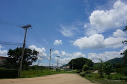 Power lines stretch across a clear blue sky with fluffy white clouds. Below, a rural area features green trees, grass, and a dirt path. Buildings are visible in the background, and distant hills rise toward the horizon.