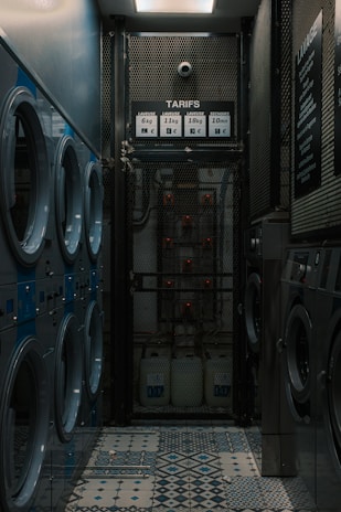 A narrow room contains several rows of washing machines and dryers lined up along both walls. The machines have blue and silver accents. The floor features a tiled pattern with various geometric designs. At the far end of the room, there is a metal mesh gate with a sign displaying washing and drying prices. The lighting is dim, creating a somewhat industrial and utilitarian atmosphere.
