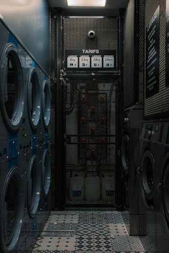 A narrow room contains several rows of washing machines and dryers lined up along both walls. The machines have blue and silver accents. The floor features a tiled pattern with various geometric designs. At the far end of the room, there is a metal mesh gate with a sign displaying washing and drying prices. The lighting is dim, creating a somewhat industrial and utilitarian atmosphere.