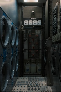 A narrow room contains several rows of washing machines and dryers lined up along both walls. The machines have blue and silver accents. The floor features a tiled pattern with various geometric designs. At the far end of the room, there is a metal mesh gate with a sign displaying washing and drying prices. The lighting is dim, creating a somewhat industrial and utilitarian atmosphere.