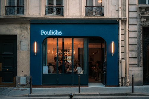 A warmly lit restaurant with large windows, situated in a historical building. The exterior features a blue facade with a glowing sign reading 'Pouliche'. Inside, people are seated at tables, and the lighting fixtures add an inviting atmosphere.