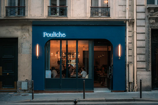 A warmly lit restaurant with large windows, situated in a historical building. The exterior features a blue facade with a glowing sign reading 'Pouliche'. Inside, people are seated at tables, and the lighting fixtures add an inviting atmosphere.