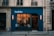 A warmly lit restaurant with large windows, situated in a historical building. The exterior features a blue facade with a glowing sign reading 'Pouliche'. Inside, people are seated at tables, and the lighting fixtures add an inviting atmosphere.