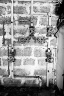 A black and white image showing a system of old, rusty pipes and valves mounted on a rough brick wall. The scene is industrial and appears to be part of a plumbing system. The pipes are arranged horizontally and vertically with several turning wheels for controlling the flow.