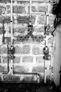 A black and white image showing a system of old, rusty pipes and valves mounted on a rough brick wall. The scene is industrial and appears to be part of a plumbing system. The pipes are arranged horizontally and vertically with several turning wheels for controlling the flow.