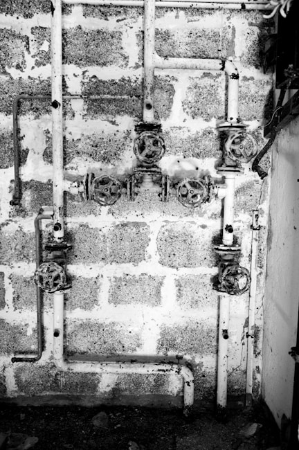 A black and white image showing a system of old, rusty pipes and valves mounted on a rough brick wall. The scene is industrial and appears to be part of a plumbing system. The pipes are arranged horizontally and vertically with several turning wheels for controlling the flow.