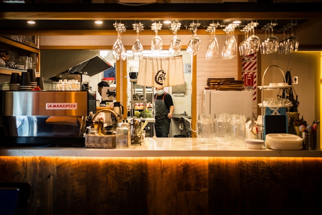 A cozy and neatly organized coffee shop interior with a bar area featuring glasses hanging upside down. There's a large espresso machine on the counter alongside various condiments and utensils. Behind the counter, a person is seen working in the kitchen area, which is partially visible through an opening adorned with a curtain.