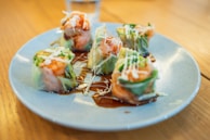 A plate neatly arranged with colorful sushi rolls featuring salmon and fresh greens wrapped in rice paper, drizzled with a dark soy sauce and topped with thin strands of white mayonnaise. The setting is bright and inviting, with the dish taking center stage on a wooden table.