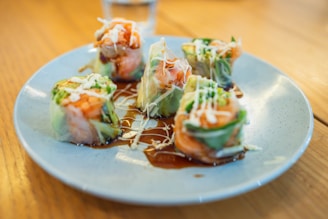 A plate neatly arranged with colorful sushi rolls featuring salmon and fresh greens wrapped in rice paper, drizzled with a dark soy sauce and topped with thin strands of white mayonnaise. The setting is bright and inviting, with the dish taking center stage on a wooden table.