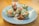 A plate neatly arranged with colorful sushi rolls featuring salmon and fresh greens wrapped in rice paper, drizzled with a dark soy sauce and topped with thin strands of white mayonnaise. The setting is bright and inviting, with the dish taking center stage on a wooden table.