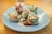 A plate neatly arranged with colorful sushi rolls featuring salmon and fresh greens wrapped in rice paper, drizzled with a dark soy sauce and topped with thin strands of white mayonnaise. The setting is bright and inviting, with the dish taking center stage on a wooden table.