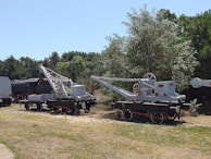 Two vintage railway cranes are placed on a grassy area surrounded by trees. The cranes have large wheels and mechanical components, and one of them is positioned on tracks alongside an old steam locomotive.