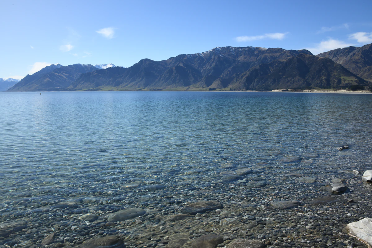 Clear water over stones at the edge of Lake Hawea, with brown mountains behind
