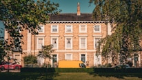A row of classic, brick townhouses is framed by trees in the foreground. The building is three stories high, with white-framed windows and a dark slate roof. A bright yellow van and a red car are parked in front of the townhouses on a sunny day.