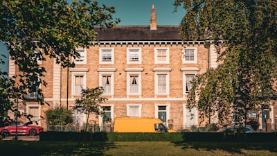 A row of classic, brick townhouses is framed by trees in the foreground. The building is three stories high, with white-framed windows and a dark slate roof. A bright yellow van and a red car are parked in front of the townhouses on a sunny day.