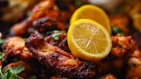 A close-up of grilled chicken wings, richly glazed and charred, garnished with two slices of lemon and sprinkled with fresh cilantro.
