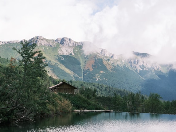 A serene mountain landscape features a wooden cabin nestled among lush greenery beside a tranquil lake. Towering mountains in the background are partially shrouded in mist, with patches of clouds hovering around the peaks, creating a peaceful and natural setting.