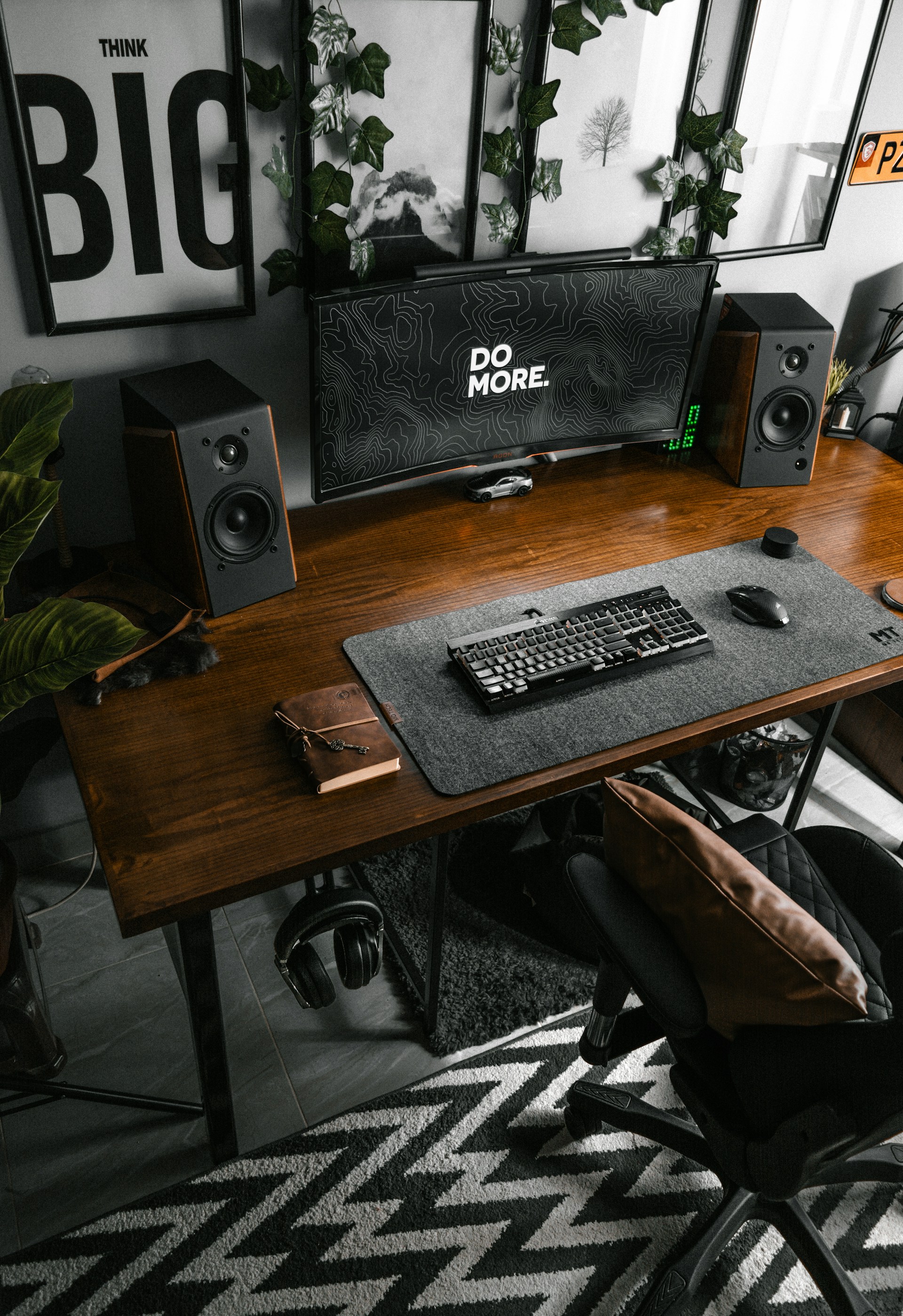A modern workstation featuring a wooden desk with a large monitor displaying the words 'DO MORE'. Two speakers are positioned on either side of the monitor. The desk also holds a keyboard and a book or notebook. Plants and framed artworks decorate the wall behind the setup. A comfortable office chair with a cushion is placed in front of the desk, and a patterned rug lies on the floor.