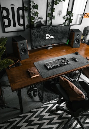 A modern workstation featuring a wooden desk with a large monitor displaying the words 'DO MORE'. Two speakers are positioned on either side of the monitor. The desk also holds a keyboard and a book or notebook. Plants and framed artworks decorate the wall behind the setup. A comfortable office chair with a cushion is placed in front of the desk, and a patterned rug lies on the floor.
