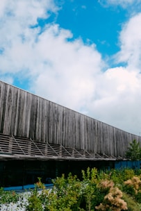 A modern building with a wooden facade and large glass windows is surrounded by lush green plants and shrubs. The sky overhead is bright blue with scattered white clouds, creating a vivid contrast with the building's natural materials.